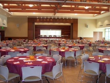 Large Event center with stage. Stages with 10' round tables covered with maroon tablecloths and  8 chairs around the tables.
